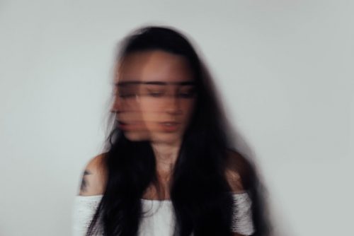 Ansiedade: a imagem mostra uma rosto feminino distorcido, como se em movimento. O rosto é de uma mulher branca de cabelos escuros e longos, que usa uma camisa branca que deixa os ombros de fora.